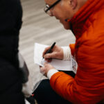 A person in a red jacket writing something on a piece of paper.