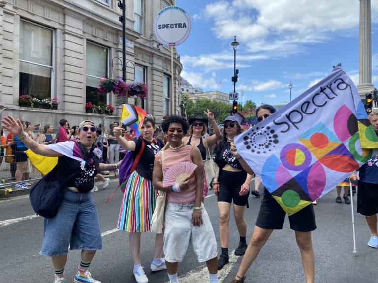 A people people posing with a Spectra colourful flag amongst many other people at a march, with many of them holding the LGBT+ flag