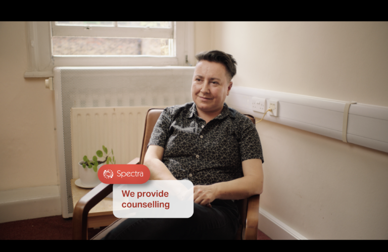 A person sitting on a chair looking indirectly towards the camera, with a small banner overlaid on the image saying: 'Spectra: We offer counselling'