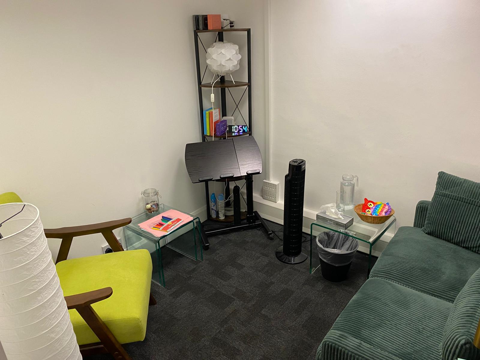 A small white room with a dark green sofa and a light green armchair, with a fan, fidget toys and a lamp.