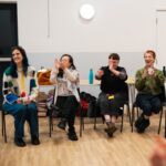 A group of people sitting in plastics chairs indoors, clapping.