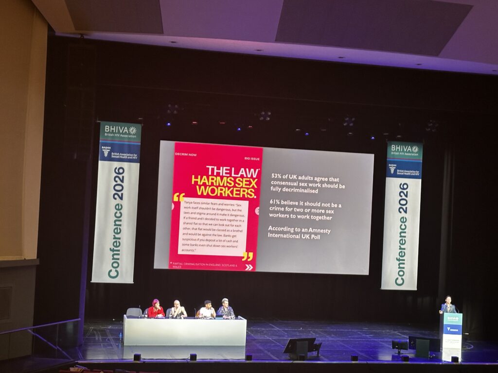 A person talking behind a podium on a stage, in front of a big screen projection that says 'The law harms sex workers'
