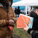 A person in a hoodie whose face we can't see talking to a person whose hands and back we can see, who is also holding some Spectra flyers.