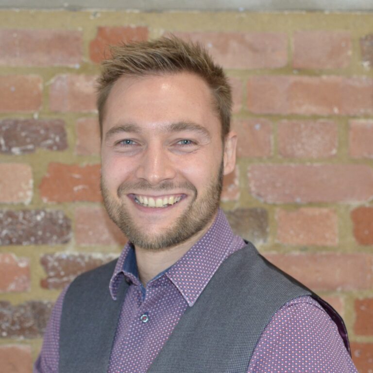 Portrait of a man with short blonde hair smiling to the camera, while standing in front of a brick wall.