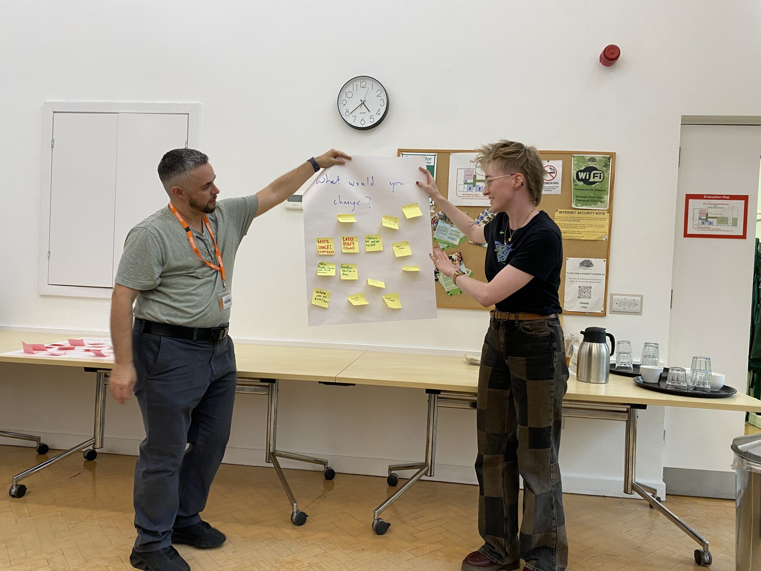 Two people standing up holding a big paper with many little yellow post-it notes on it, showing something on the big paper.