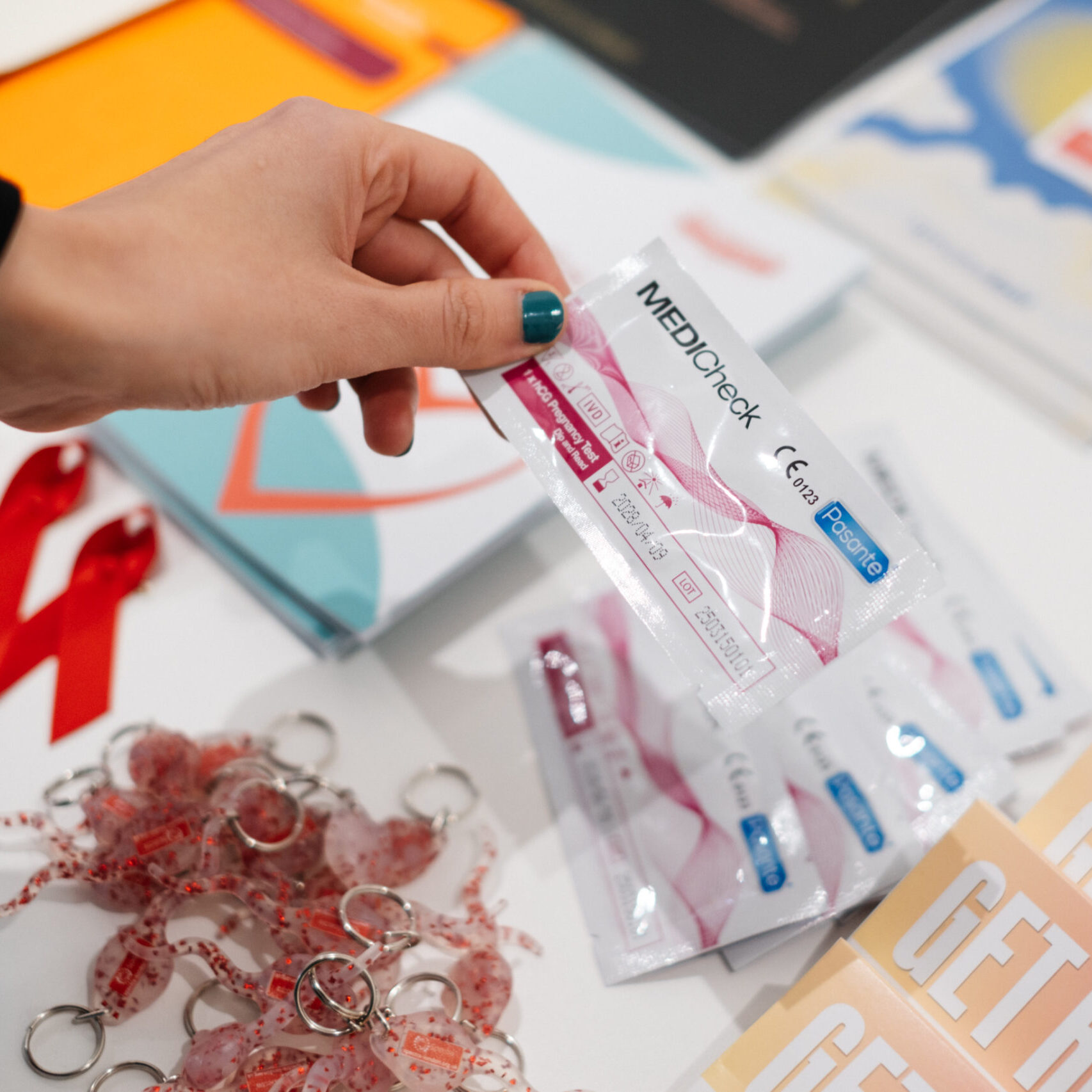 A hand holding a pregnancy test above a table with various leaflets, sperm keyrings and red HIV ribbons.